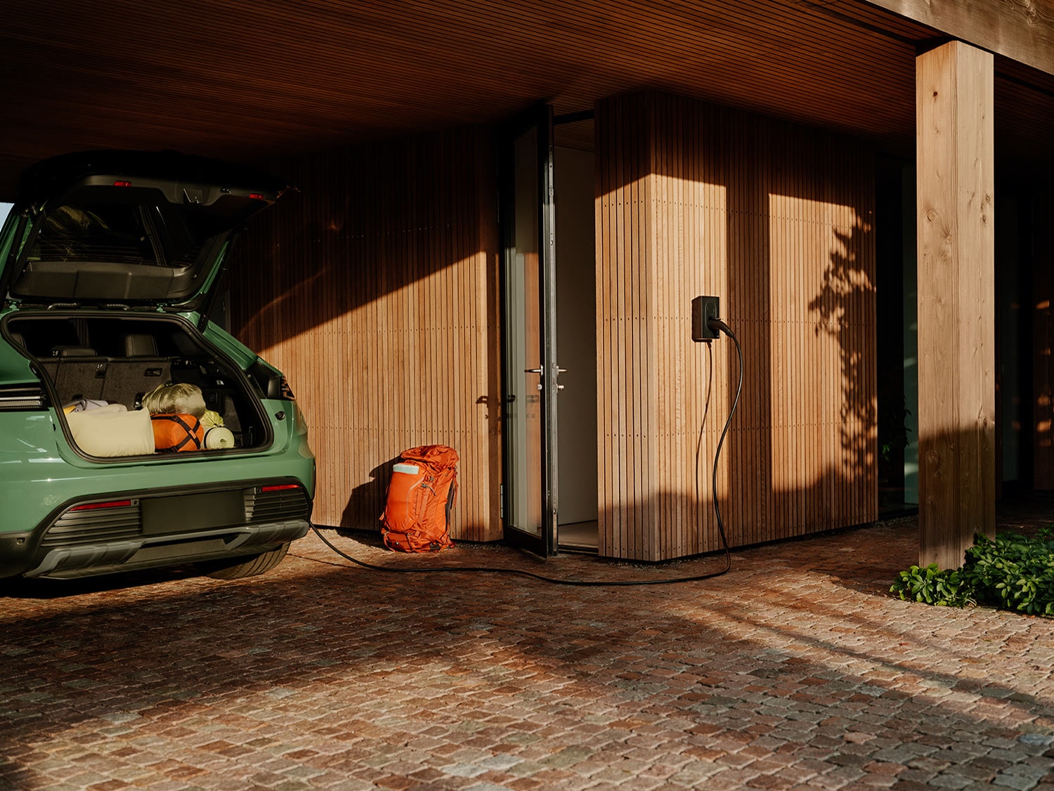 Carregador de parede numa garagem coberta de casa, ligado a um carro elétrico com o porta-malas aberto